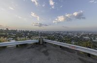 a parking lot with a concrete bench and metal railings over looking the city below a big blue cloudy sky