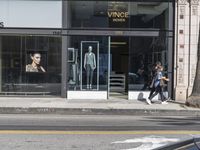 people walk past a building with windows displaying clothing stores and dresses on the sidewalk and car