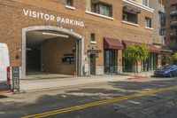 there are cars driving past this brick building in the city street way, and on the sidewalk, there is an outside building for a visitor parking area with a van