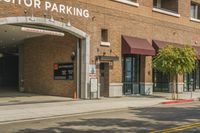 there are cars driving past this brick building in the city street way, and on the sidewalk, there is an outside building for a visitor parking area with a van