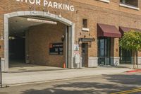 there are cars driving past this brick building in the city street way, and on the sidewalk, there is an outside building for a visitor parking area with a van