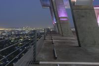 a walkway near buildings with railings looking over a city skyline at night lights shine brightly on the rooftop