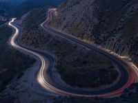 many lanes of curved road on the side of a mountain side at dusk with cars going along it