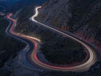 many lanes of curved road on the side of a mountain side at dusk with cars going along it