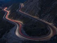 many lanes of curved road on the side of a mountain side at dusk with cars going along it