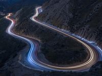 many lanes of curved road on the side of a mountain side at dusk with cars going along it