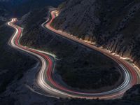 many lanes of curved road on the side of a mountain side at dusk with cars going along it