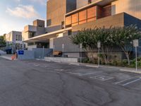 some people are walking in an empty parking lot near buildings with trees and a few concrete blocks