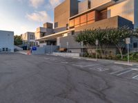 some people are walking in an empty parking lot near buildings with trees and a few concrete blocks