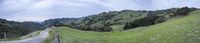 view of the side of a winding road near mountains with trees in the distance, and hills covered in grass