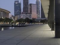 the walkway at the park leads to a lake and buildings in the background as evening falls