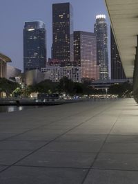 the walkway at the park leads to a lake and buildings in the background as evening falls