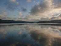 a calm lake with some clouds on it and a tree in the distance with the sky reflected in the water