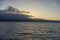 the water in front of a mountain is very calm at sunset with low clouds above