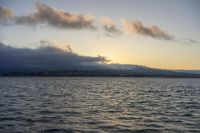 the water in front of a mountain is very calm at sunset with low clouds above