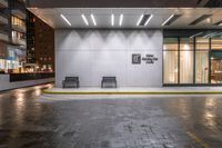 two benches outside of an office building in the rain at night time with a door open on both sides