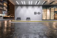 two benches outside of an office building in the rain at night time with a door open on both sides