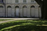 an empty park bench on the grass under a tree and large stone building with arched windows