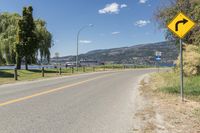 an empty street has a yellow sign with a left turn ahead next to a scenic lake and trees