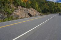 a motorcycle driving down a highway in the woods with trees and rocks on the hillside