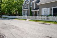 a white fence in front of some grey houses and trees on the street next to a curb