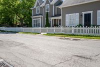 a white fence in front of some grey houses and trees on the street next to a curb