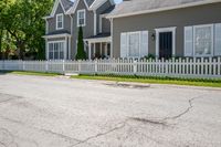 a white fence in front of some grey houses and trees on the street next to a curb