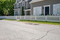 a white fence in front of some grey houses and trees on the street next to a curb