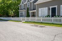 a white fence in front of some grey houses and trees on the street next to a curb