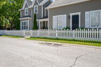 a white fence in front of some grey houses and trees on the street next to a curb