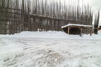 a large wooden structure covered with snow by a tree line fence with a cow in the background