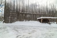 a large wooden structure covered with snow by a tree line fence with a cow in the background