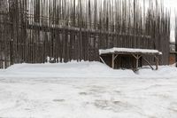 a large wooden structure covered with snow by a tree line fence with a cow in the background