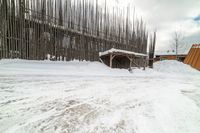 an empty wooden structure surrounded by snow on the ground and building in the background,