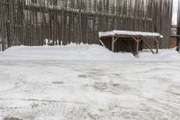 snow covered area in front of wooden structure and building in distance, with one snowy bench in the middle of it