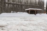 snow covered area in front of wooden structure and building in distance, with one snowy bench in the middle of it