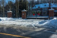 snow covered street with a driveway and gate, trees and house in background are visible