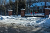 snow covered street with a driveway and gate, trees and house in background are visible