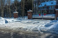 snow covered street with a driveway and gate, trees and house in background are visible