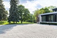 the driveway of this home has a grid pattern in it and trees, hedges, bushes and buildings behind