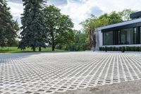 the driveway of this home has a grid pattern in it and trees, hedges, bushes and buildings behind