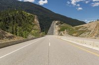 an empty open road with mountains behind it and a valley in the background, with some trees, bushes, and blue sky