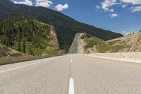 an empty open road with mountains behind it and a valley in the background, with some trees, bushes, and blue sky