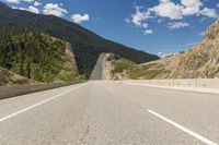 an empty open road with mountains behind it and a valley in the background, with some trees, bushes, and blue sky