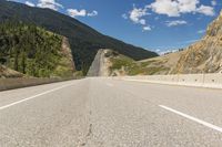 an empty open road with mountains behind it and a valley in the background, with some trees, bushes, and blue sky