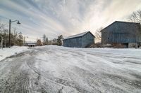 a snow covered road with two wooden barns in the distance and the sky in the background