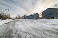 a snow covered road with two wooden barns in the distance and the sky in the background