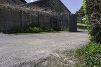 an empty driveway leads to two large barn buildings on one side and a fenced in yard behind it