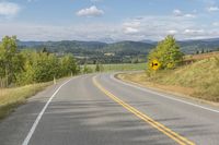 there is a yellow and black road sign on a hill side with mountains in the background