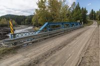 a metal bridge crosses over a dirt road near trees and a river in a remote region